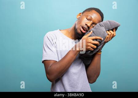 Exhausted male model holding a pillow, appearing drowsy and struggling to stay awake against a blue backdrop. Tired african american individual with eyes closed and mouth open, leaning on a cushion. Stock Photo