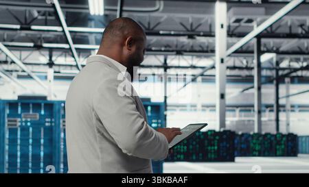 Cheerful server room engineer troubleshooting backend operations using software on tablet. Jolly data center programmer compiling software patches to fix bugs using device, camera B Stock Photo