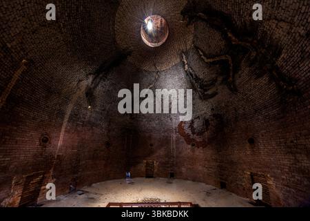 The interior of the Four des Casseaux porcelain kiln in the Four des Casseaux museum (Musée du Four des Casseaux) on Rue Donzelot in Limoges, Haute-Vienne, Nouvelle-Aquitaine, France, forms a brick dome. Light falls through a round opening at the top. Dark traces and cracks run across the brickwork, and small round openings and hatches pierce the wall. A pale floor lies below, with a red railing in the foreground. The Four des Casseaux is a so-called round kiln with a reverse flame (four à flamme renversée), unique in the country. It measures 20 metres in height and 8 metres in diameter. Eight Stock Photo