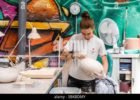 At the Ateliers Arquié art porcelain workshop on Avenue Baudin in Limoges, Haute-Vienne, Nouvelle-Aquitaine, France, a craftswoman smooths a large, egg-shaped porcelain body with a sponge. She wears disposable gloves and a white T-shirt with a small red heart, and her forearm is tattooed. A wall clock and a fan hang behind her in front of graffiti in green, orange and purple. The workshop was founded in 1996 and specialises in the design and production of customisable and artistically designed porcelain objects. It combines traditional craftsmanship with modern shapes, colours and techniques.  Stock Photo