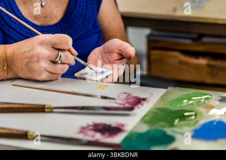 A porcelain painter hand-paints a small porcelain plaque with a fine brush in the Ateliers Arquié art porcelain workshop on Avenue Baudin in Limoges, Haute-Vienne, Nouvelle-Aquitaine, France. She holds the piece in her left hand, and a colourful motif with a blue edge is taking shape on the plaque. Brushes and a palette with green, blue and purple paint lie in front of her. She wears a blue top, a gold bracelet and rings. The workshop was founded in 1996 and specialises in the design and production of customisable and artistically designed porcelain objects. It combines traditional craftsmansh Stock Photo
