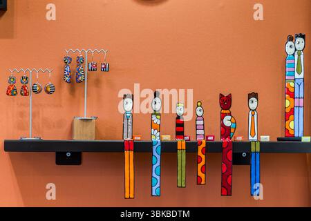 Enamel figures by the artist Delphine Peytour-Quendolo stand on a black wall shelf in the boutique "Le Bocal" on Boulevard Louis Blanc in Limoges, Haute-Vienne, Nouvelle-Aquitaine, France. The slender, colourful figures with large eyes let long, patterned legs dangle over the edge, among them a red cat and a couple. Earrings hang on stands on the left. The artist works with enamel on copper. Her figures feature bold colours, simple geometric shapes and striking eyes. Enamel has a long and significant tradition in the city. As early as the Middle Ages, from the 12th century onwards, an importan Stock Photo