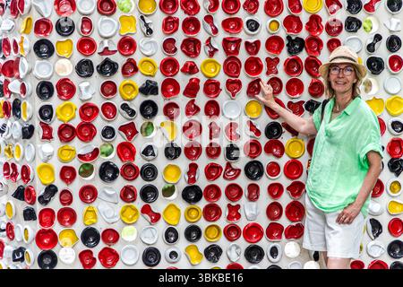 The art installation "Une Suite" by the artists Haguiko and Jean-Pierre Viot fills the frame with red, yellow, black and white porcelain bowls in front of the Musée National Adrien Dubouché on Place Winston Churchill in Limoges, Haute-Vienne, Nouvelle-Aquitaine, France. Journalist Angela Berg, wearing a straw hat, glasses and a green blouse, stands in front of it, smiling and pointing at a red bowl. The work consists of coloured, partly deformed porcelain bowls suspended from a tall, white concrete stele. In total, it comprises around 600 porcelain bowls in various colours and shapes. The muse Stock Photo