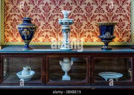 Three ornamental porcelain vases stand on a dark wooden cabinet against a wall with a red ornamental pattern in the Musée National Adrien Dubouché on Place Winston Churchill in Limoges, Haute-Vienne, Nouvelle-Aquitaine, France. On the left is a cobalt-blue lidded vase with gold decoration and handles, in the centre a white-and-blue vase on a column base, and on the right a cobalt-blue chalice-shaped vase with a gold shell motif. The cabinet's glass compartments hold a white washbasin with a jug, a white vase with handles and a white footed plate. The museum was founded in 1845 as the city's fi Stock Photo