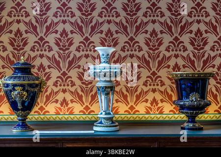 Three ornamental porcelain vases stand against a wall with a red ornamental pattern and a yellow frieze in the Musée National Adrien Dubouché on Place Winston Churchill in Limoges, Haute-Vienne, Nouvelle-Aquitaine, France. On the left is a cobalt-blue lidded vase with gold decoration, handles and a golden coat-of-arms medallion. In the centre stands a white-and-blue vase on a column base, and on the right a cobalt-blue chalice-shaped vase with a gold shell motif. The museum was founded in 1845 as the city's first museum. Adrien Dubouché, a patron of the arts and son of a cloth merchant, took o Stock Photo