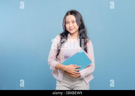 A woman is holding a book and a backpack. She is smiling and looking at the camera. Concept of confidence and readiness, as the woman is prepared for Stock Photo
