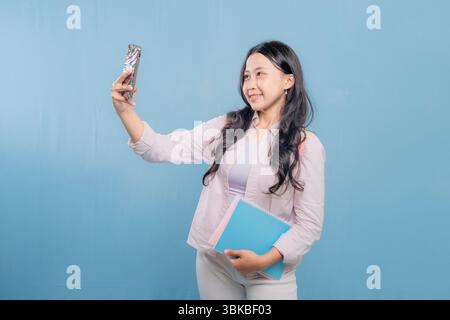A woman is taking a selfie with her cell phone. She is wearing a pink shirt and blue pants. She is holding a blue book and a pink book Stock Photo