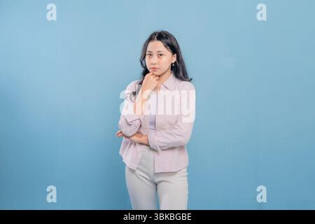 A woman is standing in front of a blue wall, wearing a pink jacket and white pants. She is looking down and she is deep in thought Stock Photo