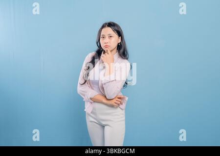 A woman is standing in front of a blue wall, wearing a pink jacket and white pants. She is looking down and she is deep in thought Stock Photo