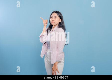 A woman is standing in front of a blue wall, wearing a pink jacket and white pants. She is looking down and she is deep in thought Stock Photo