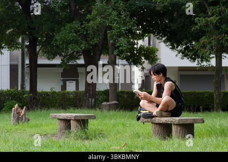 A young Japanese man sitting on a small park bench reading a book in rural Kanagawa, Japan. Stock Photo