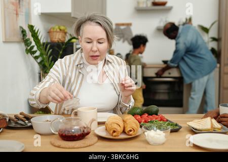 Family enjoying breakfast together in kitchen. Diverse array of food items such as croissants, avocados, tea, and cereal present on the table Stock Photo