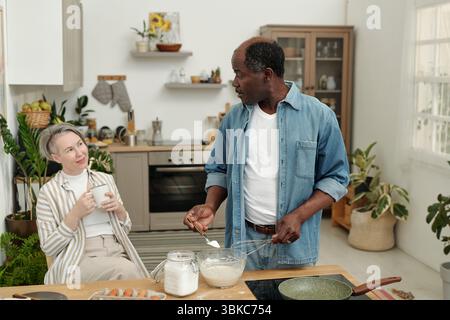 Multiethnic couple preparing breakfast in modern kitchen, surrounded by plants and cooking utensils, sharing moment of conversation and laughter Stock Photo