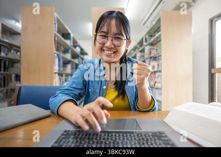 A happy Asian university student smiles and raises her fist in celebration, feeling successful after completing an online assignment or exam on her la Stock Photo