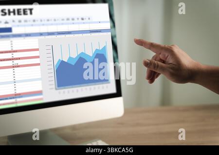 A man working on excel person pointing at computer screen displaying spreadsheet and line graph, analyzing data and trends in professional setting Stock Photo