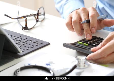Close-up of a person using a calculator with a pen in hand, alongside a tablet with a keyboard, eyeglasses, and a stethoscope on the desk. This image Stock Photo