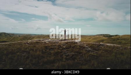 Back view of a young female tourist standing with raised arms on top of a hill and enjoying the beautiful landscape of green hills under a blue cloudy sky Stock Photo