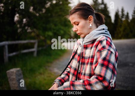 Woman wearing plaid jacket and hoodie stands outdoors in a quiet, wooded area, looking down thoughtfully. Stock Photo