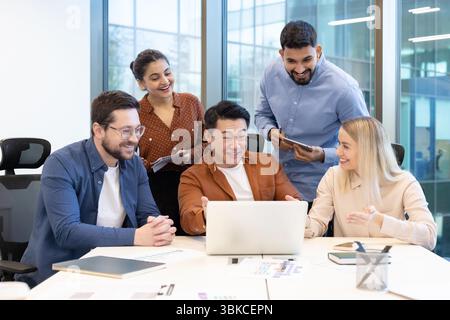 A diverse team collaborates around a laptop in a bright office environment, showcasing teamwork and communication. Stock Photo
