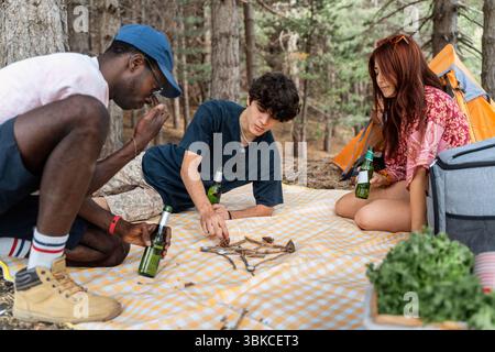 A group of diverse young friends sit on a blanket, playing a creative game with sticks and pinecones during a camping trip in the woods Stock Photo