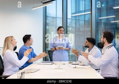 A diverse medical team applauds in a modern office setting. A female doctor stands, holding a tablet and smiling. Stock Photo