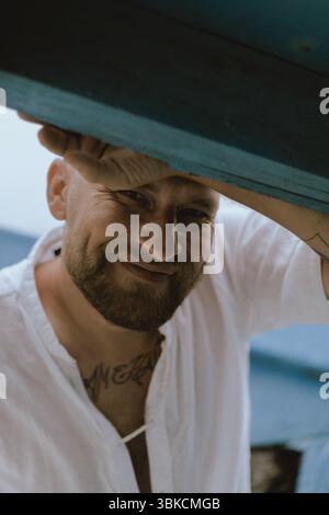 Smiling bearded man in white shirt leaning under blue wooden beams Stock Photo