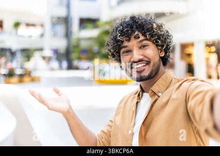 A smiling man with curly hair takes a selfie outside, extending his arm towards the viewer, invitingly. Stock Photo