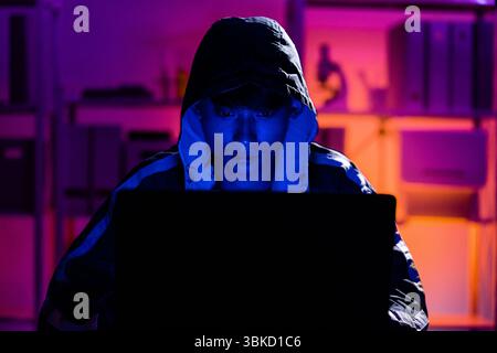 A young man in a black hoodie intently using a laptop in a neon-lit room, evoking themes of cybersecurity, hacking, coding, and digital privacy in a t Stock Photo