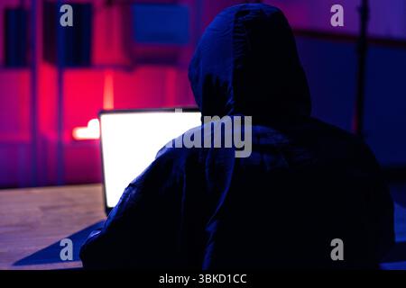 A young man in a black hoodie intently using a laptop in a neon-lit room, evoking themes of cybersecurity, hacking, coding, and digital privacy in a t Stock Photo