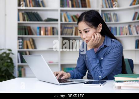 A focused Asian woman works on a laptop in a library, surrounded by books and a digital tablet. Stock Photo