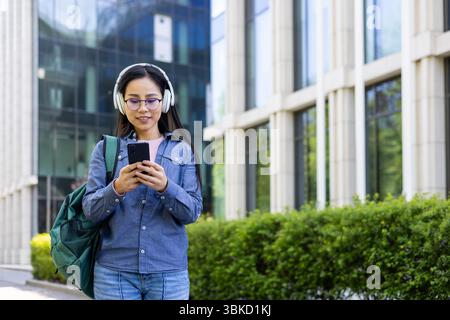 A young Asian woman in glasses and headphones looks at her phone while walking outside in an urban setting. Stock Photo