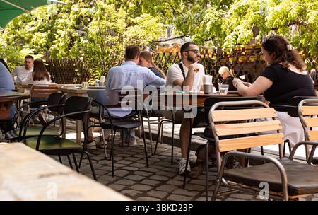 People are seen enjoying their time at an outdoor cafe on a sunny day. Patrons are engaged in conversation, sipping drinks and enjoying light snacks while surrounded by greenery. Despite frequent air raid sirens and the ever-present threat, the city has transformed into a vital humanitarian hub. It actively provides shelter and assistance to countless internally displaced people from more heavily affected regions, while also serving as a crucial point for collecting aid and supporting the Armed Forces. Cafes, restaurants and cultural institutions remain open though their operations are often i Stock Photo
