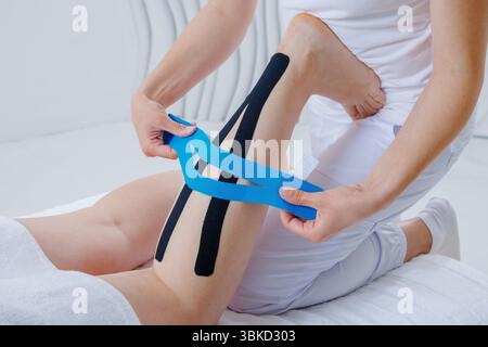 Closeup of therapist applying kinesiology tape on patients leg to reduce strain, improve mobility, and support recovery from muscle injury. Stock Photo