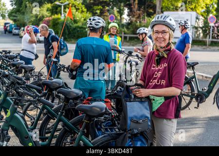 Participants in the guided cycling tour by Die Landpartie get themselves and their bikes ready in the morning for the next stage of the journey in Le Chaffaud Ouest near Saint-Martin-de-Ré on the island of Ré (Île de Ré), Charente-Maritime, Nouvelle-Aquitaine, France. Journalist Angela Berg, wearing a helmet, glasses and a neckerchief, stands in the front right and holds her blue pannier on the e-bike. Her burgundy T-shirt reads "Coup de cœur". In English: "Love at first sight". Tour guide Katell Nicoux, in a high-visibility vest and a light green helmet, stands in the background. Green e-bike Stock Photo