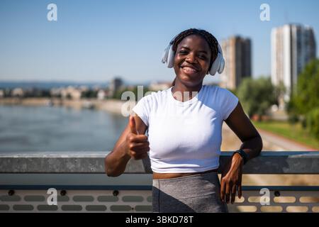 Portrait of confident sporty African-American woman in sportswear wearing headphones and showing thumb up while standing at the city bridge. Stock Photo