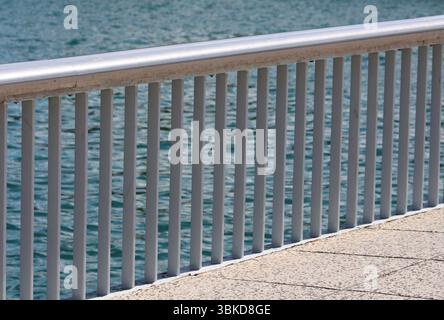 Silver metal railing with vertical bars runs along a paved waterfront, overlooking rippling blue water in bright sunlight, providing safety and a clea Stock Photo