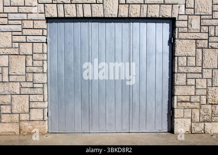 Large silver metal garage door with vertical panels is set into a textured stone wall facade, showing geometric patterns and strong sunlight casting s Stock Photo