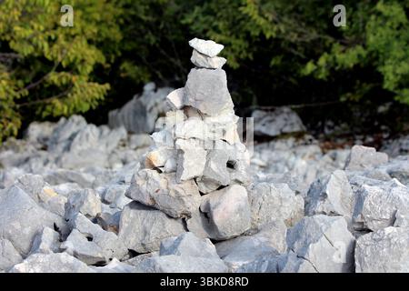 Rough stone cairn built from stacked limestone rocks stands on a rocky slope, surrounded by scattered stones with dense green forest in the background Stock Photo