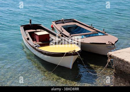 Two wooden rowboats, one with a yellow deck, float side by side in transparent blue water, tied to a concrete pier, showing reflections and details of Stock Photo