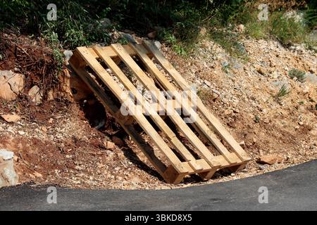 Light wooden pallet leans against a rocky dirt slope beside a new asphalt road, surrounded by gravel, stones, and sparse vegetation in a sunny roadsid Stock Photo