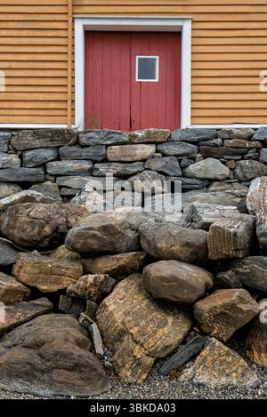 A weathered stone wall made of various rocks stands in front of an orange barn featuring a bold red door under overcast skies. Stock Photo