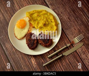 A large plate with a fried egg, mashed potatoes and two meat patties on a wooden table, cutlery nearby. Top view, flat lay. Stock Photo