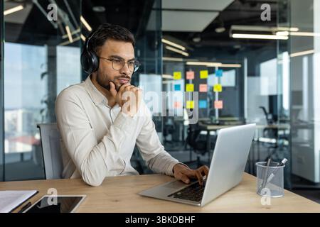 A thoughtful businessman wearing glasses and headphones, working on a laptop in a modern office environment. He is deep in concentration. Stock Photo
