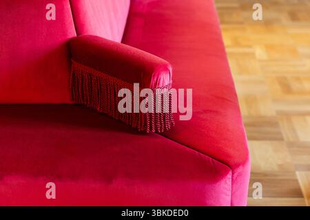 Elegant close-up of a luxurious red velvet sofa armrest adorned with decorative fringe, set against a parquet flooring background. Perfect for concept Stock Photo