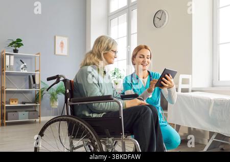 Young friendly female nurse using modern tablet with senior woman with disability in wheelchair Stock Photo