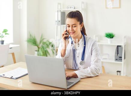 Young doctor, nurse, medical practitioner talking to patient on mobile phone, smiling, using laptop  Stock Photo