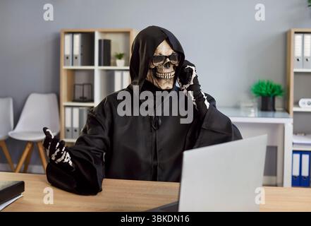 Man in death costume with black hood and skeleton mask sitting in office talking by mobile phone Stock Photo