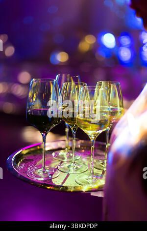 A server's tray holding multiple wine glasses with different coloured beverages, set against a blurry, purple-lit background Stock Photo