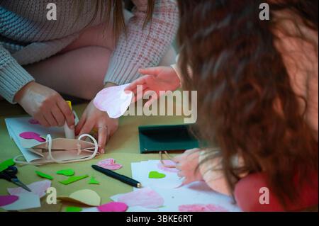 Two individuals collaborate on creating handmade cards, using paper hearts and glue. The setting is cozy and intimate, suggesting a close bond. Stock Photo