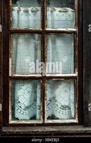 The rustic window displays delicate lace curtains with intricate floral designs, slightly dampened from outside moisture, Puy de Dome, Auvergne,France Stock Photo